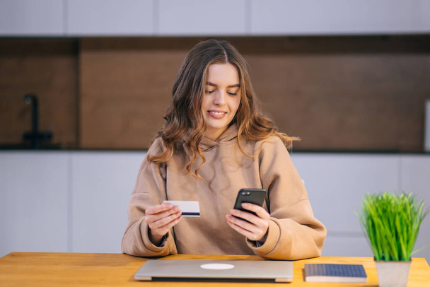 Mladá krásná žena pomocí chytrého telefonu a kreditní karty nakupovat on-line doma, zatímco sedí u stolu. - Fotografie, Obrázek