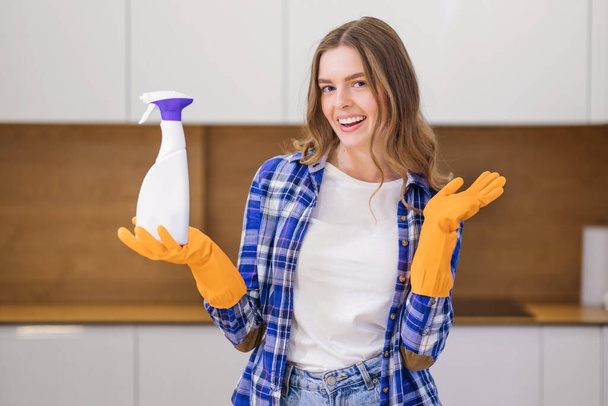 Una criada positiva sostiene un detergente químico en una camisa y una camiseta, guantes, en la cocina. - Foto, imagen
