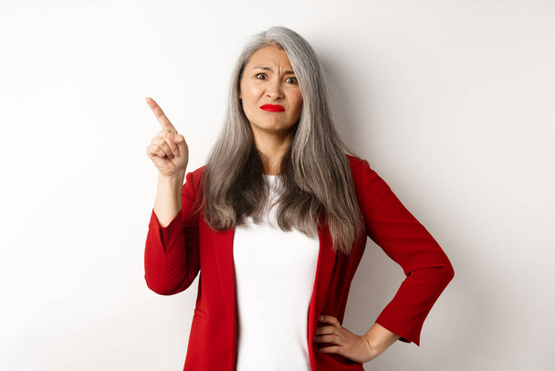 Sceptische Aziatische zakenvrouw in rode blazer grimmig, wijzen linksboven hoek en kijken ontevreden, afkeuren iets, staan over witte achtergrond - Foto, afbeelding