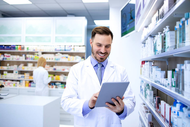 Professional handsome male pharmacist working in pharmacy store or drugstore. Checking medicines on his tablet computer. Healthcare and apothecary. - Foto, Imagen