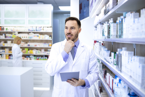 Profesjonalny przystojny aptekarz w białym płaszczu trzymający tabletkę i myślący w aptece lub aptece. Opieka zdrowotna i apteka. - Zdjęcie, obraz