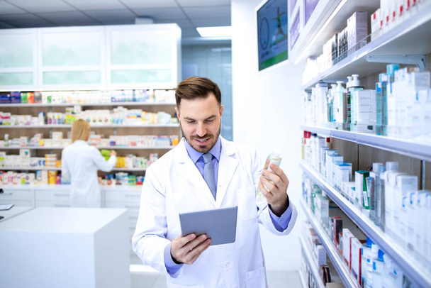 Professional handsome male pharmacist working in pharmacy store or drugstore. Checking medicines on his tablet computer. Healthcare and apothecary. - Fotografie, Obrázek