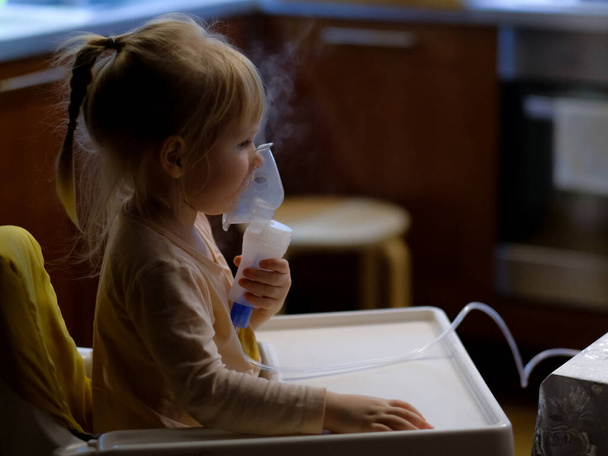 A little girl holds a mask in her face and inhales the medicine evaporated by a nebulizer. - Foto, Bild