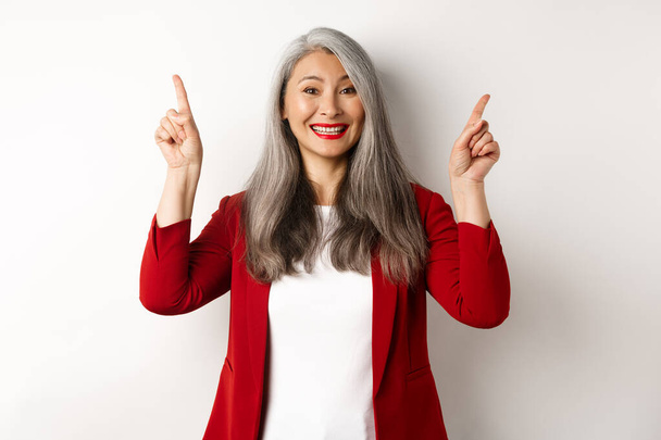 Mujer madura feliz en chaqueta roja y maquillaje, sonriendo y mostrando publicidad en la parte superior, señalando los dedos hacia arriba en el logotipo, fondo blanco - Foto, Imagen