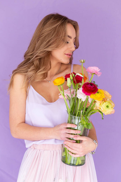 A girl in a silk dress holds a bouquet of flowers in a glass vase. Beautiful girl for Valentine's Day with a bouquet in her hands. Beautiful girl on March 8. Portrait of a girl on a purple background with flowers. - Foto, afbeelding