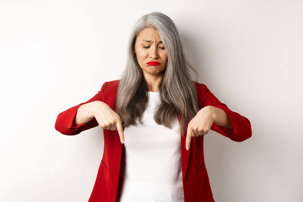 Traurige und düstere asiatische Seniorin im roten Blazer schmollt, zeigt mit dem Finger nach unten und schaut aufgebracht über weißen Hintergrund - Foto, Bild