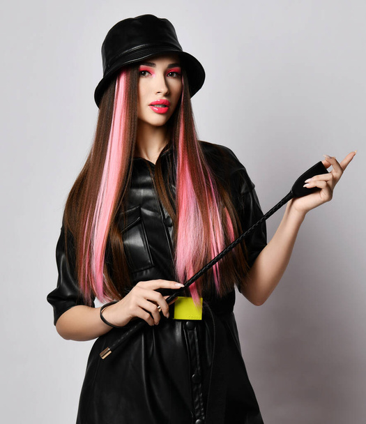Woman with colored hair and pouty lips wearing black leather dress and hat stands holding riding crop whip in hands - Foto, imagen