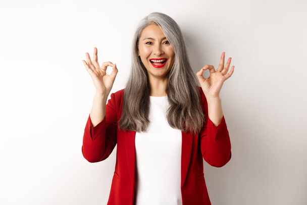 Mulher asiática sênior bonita em blazer vermelho, sorrindo e mostrando sinais ok, aprovar e dizer sim, de pé sobre fundo branco - Foto, Imagem