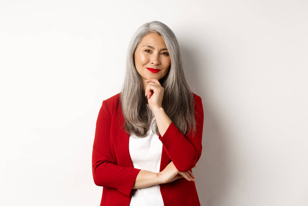 Бизнес-концепция. Азиатская зрелая деловая женщина улыбается приятно, глядя вдумчиво, имея идею, стоя в красном блейзере на белом фоне - Фото, изображение