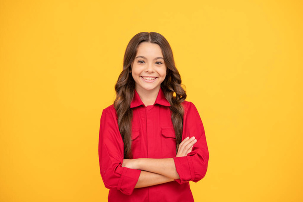 smiling kid crossed hands. happy childhood. cheerful teen girl with long curly hair. kid wear red shirt on yellow background. child express happiness. positive emotions. - Photo, Image