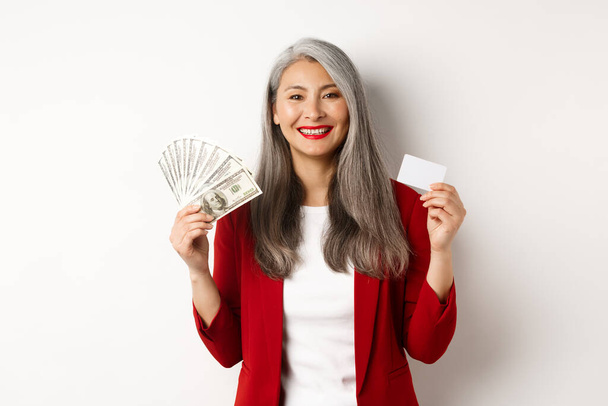 Exitosa mujer de negocios asiática senior mostrando dinero en dólares y tarjeta de plástico, sonriendo feliz a la cámara, usando chaqueta roja y maquillaje - Foto, imagen