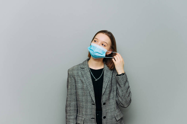 Jovem mulher de negócios fica em um fundo cinza e remove uma máscara protetora de seu rosto. - Foto, Imagem