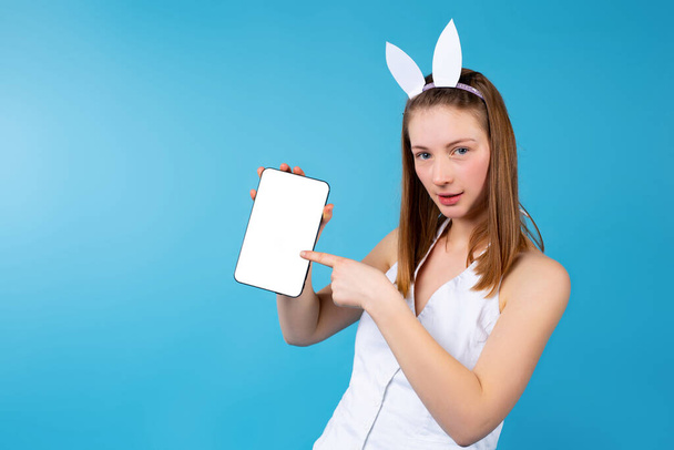 Blonde girl wearing bunny ears, pointing forefinger to empty copy space white mock up screen tablet. Female blogging, Promotional Easter on blue background with side space - Φωτογραφία, εικόνα