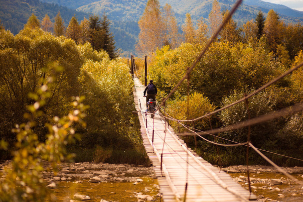 Homem irreconhecível andando com motocicleta cross country na ponte suspensa sobre o rio montanhas - Foto, Imagem