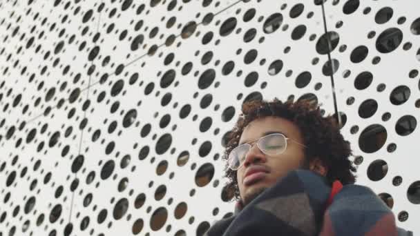 Χαμηλή γωνία του νεαρού Αφροαμερικανού άνδρα στέκεται δίπλα από διάτρητο τοίχο του αστικού κτιρίου την ημέρα του χειμώνα και κοιτάζοντας προς τα κάτω την κάμερα - Πλάνα, βίντεο