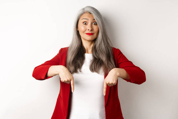 Hermosa asiática mujer mayor en elegante chaqueta roja, señalando los dedos hacia abajo y sonriendo emocionada, mostrando publicidad, fondo blanco - Foto, Imagen