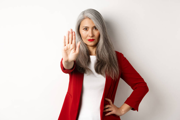 Serious asian woman in red blazer showing stop sign, telling no, frowning and rejecting something bad, standing against white background - Photo, Image