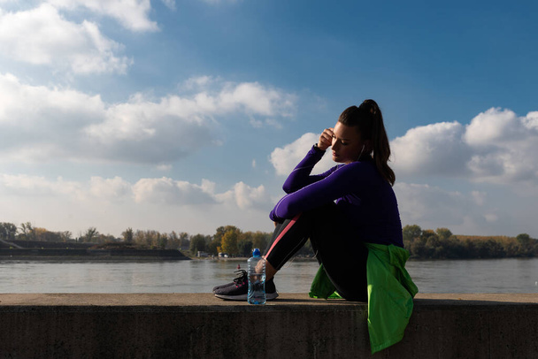 Young sweaty active fitness athletic sporty woman taking a break after outdoor exercising of running and jogging listening the music on her smartphone and enjoy the sunrise in the beautiful morning - 写真・画像