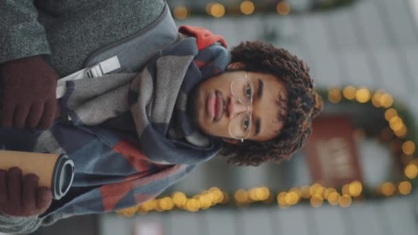 Pionowe ujęcie portretowe młodego, pewnego siebie Afro-Amerykanina z plecakiem i iść na kawę stojąc na ulicy miasta w zimowy dzień i patrząc w kamerę; światła bokeh w tle - Materiał filmowy, wideo