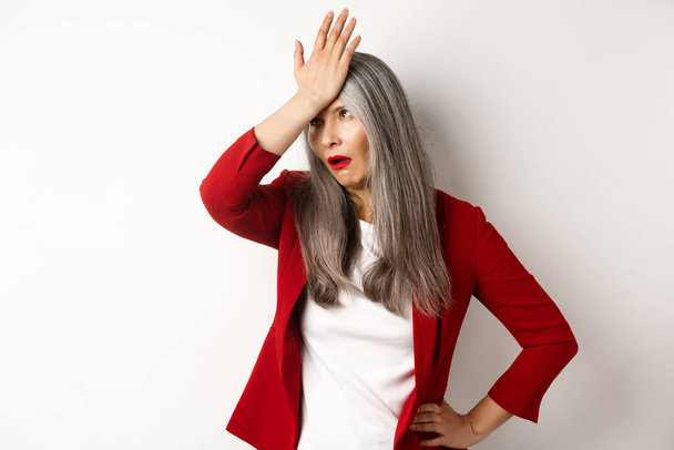 Annoyed asian office lady in red blazer roll eyes and facepalm, standing bothered and irritated over white background - Photo, Image
