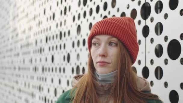 Zoom in portrait shot of young beautiful Caucasian woman in outerwear and hat standing outside of modern urban building, looking away and then posing for camera - Footage, Video