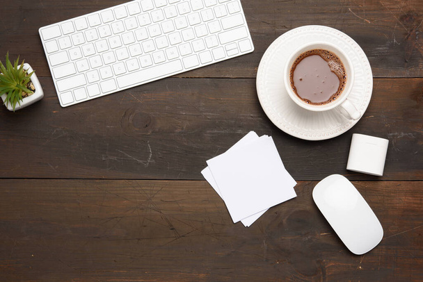 Beyaz kablosuz klavye ve kablosuz kulaklıklar ahşap kahverengi bir masada, yanında kahve ve üst görünümü olan beyaz bir bardak. - Fotoğraf, Görsel