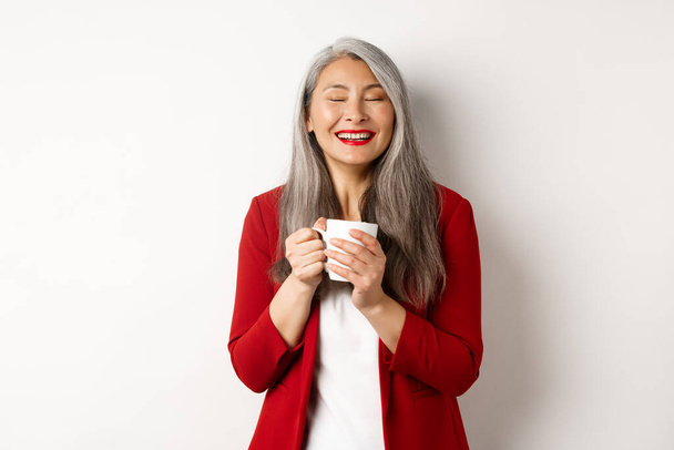 Zakenmensen concept. Happy office dame in rode blazer genieten van het drinken van koffie, met mok en glimlachen blij, staande over witte achtergrond - Foto, afbeelding