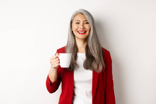 Business people concept. Happy asian businesswoman in red blazer smiling, drinking coffee from mug, standing over white background - Photo, Image