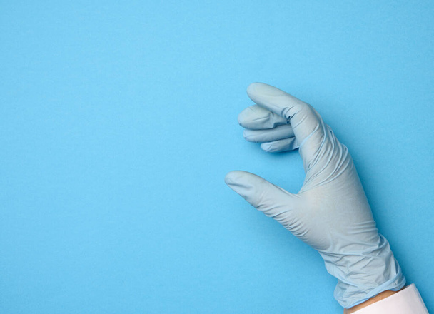 ręka żeńska w niebieskich rękawiczkach lateksowych na niebieskim tle, dłoń trzymająca przedmiot - Zdjęcie, obraz