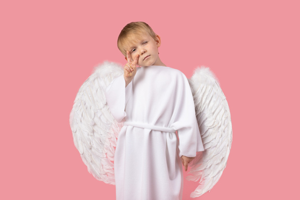 Angel with wings in white clothes stand showing selfie or peace sign looking into camera. Studio shot on pink background. Concept postcard for valentine's day or religion. - Photo, Image