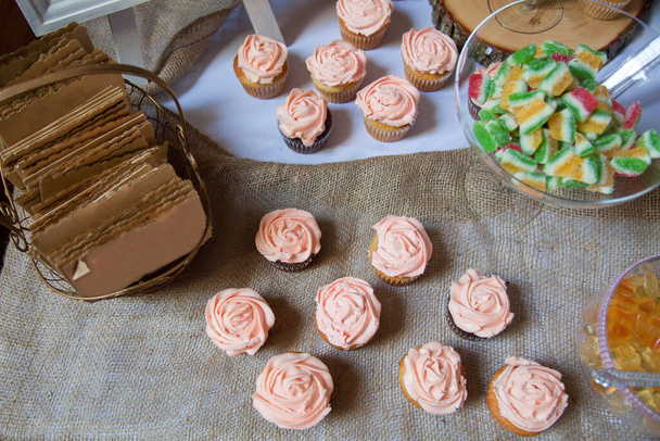 Růžové mražené vanilkové a čokoládové cupcakes sedět na ubrusu s jinými bonbóny v pozadí jako dezertní stůl pro svatbu. - Fotografie, Obrázek