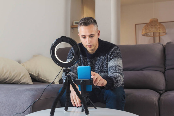 Stock photo of Young man millennial influencer sit on sofa preparing a setup with mobile phone and lighting for vlogging or video call. Social media video blog recording concept - Photo, Image