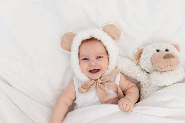 毛布の下にテディベアの耳を持つ面白い帽子の子供。子供のための織物とベッドリネン。新生児が目を覚ましたか寝ようとしている - 写真・画像