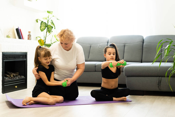 senior grandmother workout yoga withgranddaughters preschooler. They are sitting on mat at cozy home interior. Sport, parenthood and people concept - Photo, Image