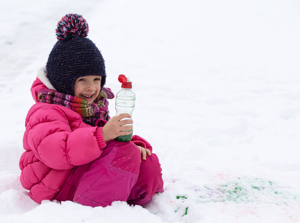 Uma garota bonita com uma garrafa de tinta desenha na neve. O conceito de criatividade infantil e diversão de inverno. - Foto, Imagem