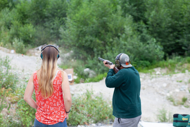 A young women stands behind a man holding a shotgun downrange, shooting clay pigeons to practice aim in British-Columbia - 写真・画像