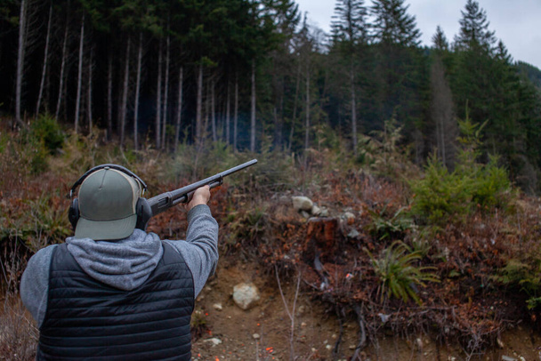 A mean wearing a vest, hat and ear protection holds an old 12 gauge shotgun, aiming and shooting at orange clay pigeons to practice. Pump action shotgun with a wooden stock. - Foto, imagen