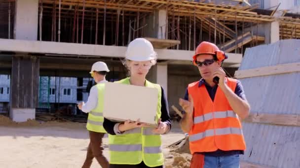 In front of the camera walking engineer lady and foreman man with a protective helmets and goggles they analyzing the plan of construction man using ration to have a discussing with workers from the - Footage, Video