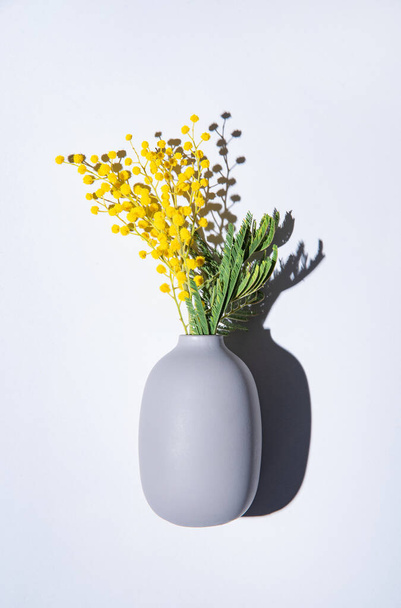 un ramo de flores de mimosa amarilla se encuentra en un jarrón de cerámica con sombra sobre un fondo gris. concepto del 8 de marzo, feliz día de la mujer. vista superior y espacio de copia - Foto, Imagen