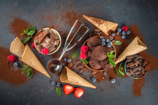 Тёмное шоколадное мороженое. Много шоколадного мороженого с шоколадной фасолью, порошком и кусочками, с вафельными рожками из мороженого и летними свежими ягодами на темно-бетонном фоне - Фото, изображение
