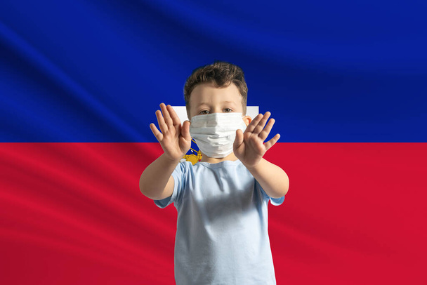 Kleine blanke jongen met een beschermend masker op de achtergrond van de vlag van Haïti. Maakt een stopbord met zijn handen, thuisblijven Haïti. - Foto, afbeelding