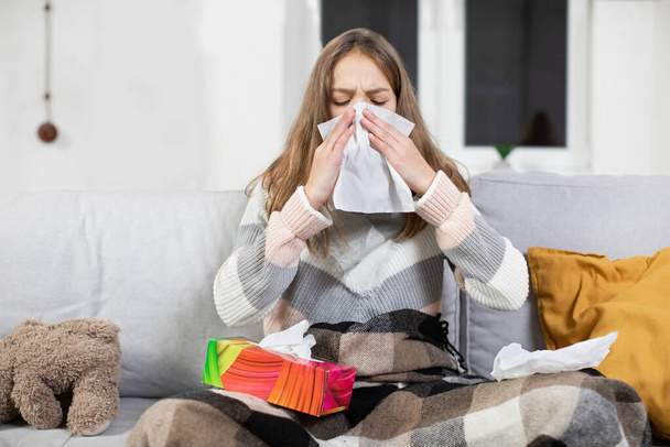 Sick tired teenager girl with scarf around neck, blowing running nose into paper handkerchief while sitting on soft sofa covered with blanket, during virus or cold, influenza disease - Photo, Image