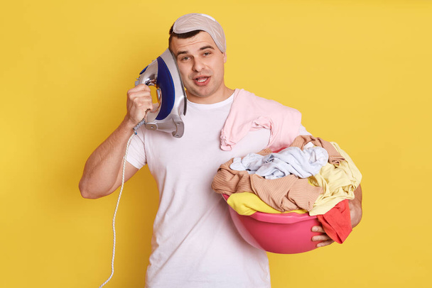 Tired bored young houseman househusband with iron near ear, being very tired, imagines he talking phone, need ironing clothes, isolated over yellow background, looks crazy. - Foto, immagini