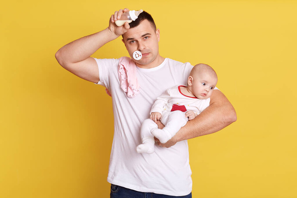Young Caucasian father feeding newborn baby from bottle milk. Man parent holding rocking daughter. looks tired, keeping arm on forehead. Single dad looking after kid,posing against yellow wall. - Fotoğraf, Görsel