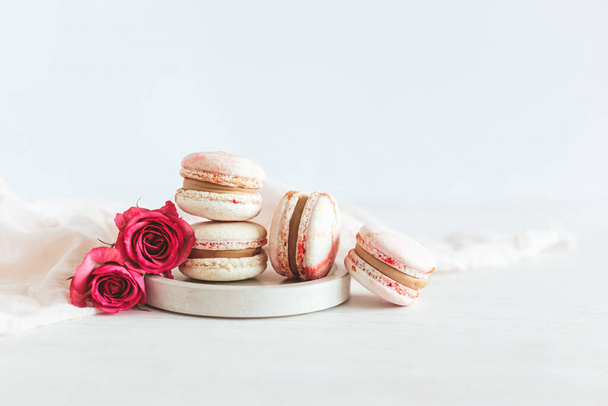 Ízletes francia macaroons rózsaszín rózsákkal a tányéron. Fehér fa háttér. A szöveg helye. - Fotó, kép