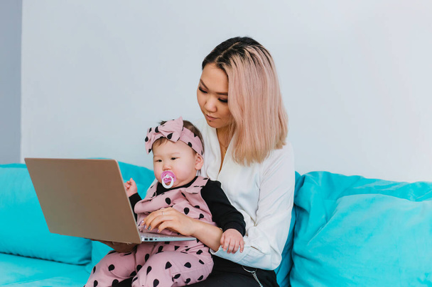 Una joven madre trabaja en Internet. Una mujer de apariencia asiática utiliza un portátil, sosteniendo a un bebé en sus brazos, sentado en un sofá azul. Trabajo desde casa - Foto, Imagen
