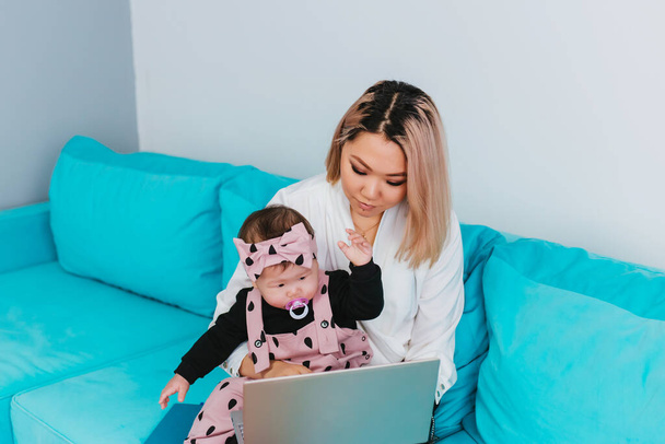 Une jeune mère travaille sur Internet. Une femme d'apparence asiatique utilise un ordinateur portable, tenant un bébé dans ses bras, assis sur un canapé bleu. Travail à domicile - Photo, image