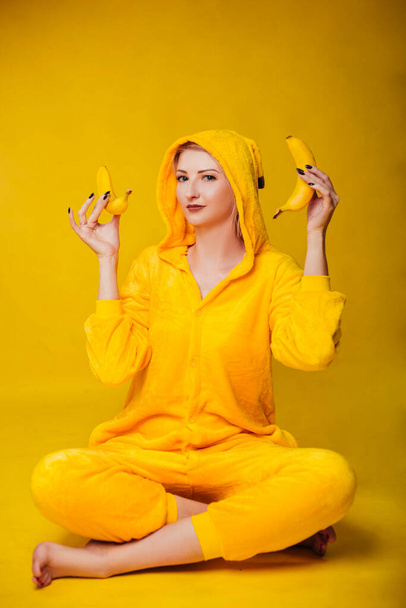 Забавная молодая женщина в жёлтой пижаме сидит на жёлтом фоне и держит в руках бананы. Веселая маленькая девочка развлекается с фруктами. Студия фото - Фото, изображение