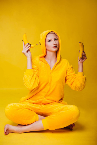 Hauska nuori nainen keltaisessa pyjamassa istuu keltaisella taustalla ja pitää banaaneja käsissään. Iloinen pikkutyttö pelleilee hedelmien kanssa. Studiokuva - Valokuva, kuva