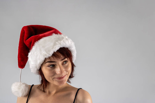 Young female attractive model with auburn hair and red lipstick in a studio with a white background wearing a sexy santa Santa hat with a black going out dress - Photo, Image